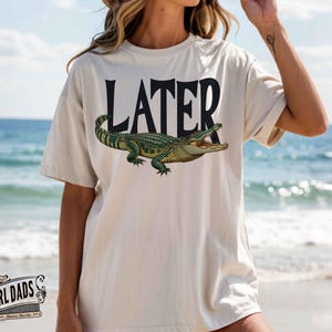 May include: Cream-colored t-shirt with the word "LATER" in black block letters above a detailed illustration of a green alligator with an open mouth. The shirt is worn on a beach.