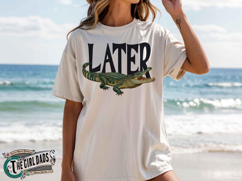 May include: Cream-colored t-shirt with the word "LATER" in black block letters above a detailed illustration of a green alligator with an open mouth. The shirt is worn on a beach.