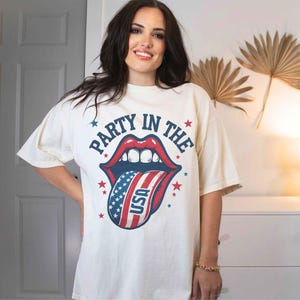K&ouml;nnte beinhalten: Ein wei&szlig;es T-Shirt mit einem Grafikdruck einer rot-wei&szlig;-blauen Zunge, die aus einem Mund herausragt. Der Text "Party in the" befindet sich &uuml;ber der Zunge und "USA" auf der Zunge.