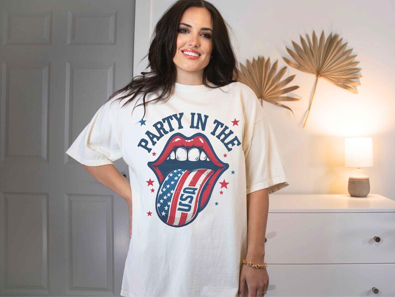 K&ouml;nnte beinhalten: Ein wei&szlig;es T-Shirt mit einem Grafikdruck einer rot-wei&szlig;-blauen Zunge, die aus einem Mund herausragt. Der Text "Party in the" befindet sich &uuml;ber der Zunge und "USA" auf der Zunge.