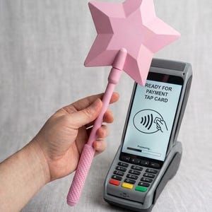 May include: A light pink star-shaped wand with a textured handle held near a gray payment terminal. The terminal screen displays the text READY FOR PAYMENT TAP CARD with a contactless payment icon and a processing bar at the bottom.