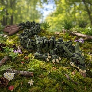 Puede incluir: Una escultura artesanal de dragón verde y negro con lunares, colocada sobre musgo con helechos, piñas y flores silvestres. El dragón tiene una cabeza detallada, garras y un cuerpo segmentado, creando una escena forestal caprichosa.