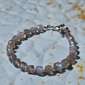 Op de afbeelding: Een armband met facetgeslepen, grijs getinte edelstenen met iriserende flitsen. De armband is geregen aan een zilverkleurige ketting en heeft een decoratieve sluiting. De edelstenen zijn ongeveer 0,8 cm in diameter.