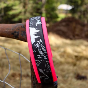 May include: A black and pink wristband with the words "Mischief Managed" and various symbols. The wristband is wrapped around a metal bar. The background is blurred, showing trees and grass.