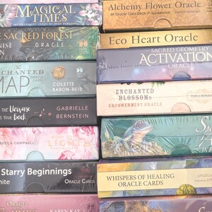 Może przedstawiać: Kolekcja talii kart wyroczni, w tym "Magical Times" i "Sacred Forest", w różnych kolorach. Pudełka są ułożone w stos, prezentując tytuły i autorów, niektóre zapakowane w folię.