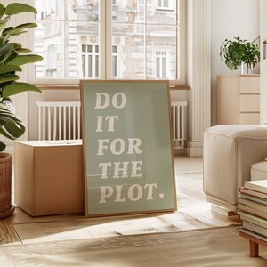 Könnte beinhalten: Ein gerahmter Druck mit hellblauem Hintergrund und weißem Text, der "DO IT FOR THE PLOT." lautet. Der Druck lehnt an einem Karton in einem Raum mit einem weißen Sofa und einem großen Fenster.