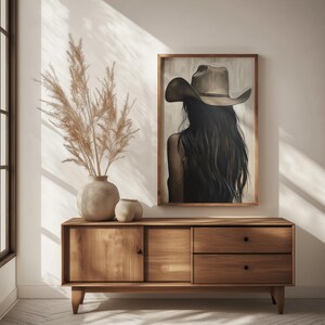 May include: A wooden dresser with two drawers and a cabinet door. A painting of a woman wearing a brown cowboy hat hangs above the dresser. A vase with dried pampas grass sits on top of the dresser.