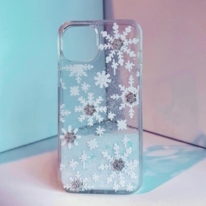 May include: A clear phone case with a glittery white background and white snowflake cutouts. The snowflakes have silver glitter accents.