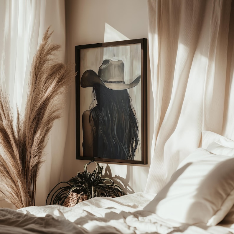 May include: A framed painting of a woman with long dark hair wearing a brown cowboy hat. The painting is hanging on a white wall above a bed with white linens.