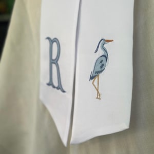 May include: White fabric with blue embroidered details. Features a large letter "R" and a heron bird design. The heron has blue feathers, a brown beak, and brown legs. The fabric is folded and appears to be part of a larger garment.