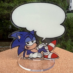 May include: A clear acrylic Sonic the Hedgehog figure with a blank speech bubble. The figure features Sonic's blue fur, white gloves, and red and white striped shoes. The base is a clear, circular shape.