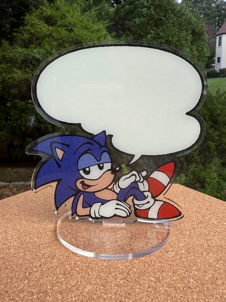 May include: A clear acrylic Sonic the Hedgehog figure with a blank speech bubble. The figure features Sonic's blue fur, white gloves, and red and white striped shoes. The base is a clear, circular shape.
