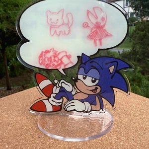 May include: A clear acrylic figurine featuring Sonic the Hedgehog, with a thought bubble above his head. The thought bubble contains pink sketches of a cat, a character with a dress, and a flower. Sonic is blue with white gloves and red and white shoes.