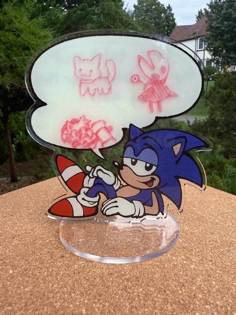 May include: A clear acrylic figurine featuring Sonic the Hedgehog, with a thought bubble above his head. The thought bubble contains pink sketches of a cat, a character with a dress, and a flower. Sonic is blue with white gloves and red and white shoes.