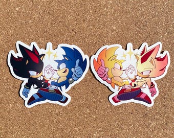 May include: Two pairs of stickers featuring the characters Sonic the Hedgehog and Shadow the Hedgehog. The stickers are in a heart shape with the characters facing each other. The first pair features Sonic and Shadow in their classic blue and black colors. The second pair features Sonic and Shadow in their Super forms, with Sonic in yellow and Shadow in red.