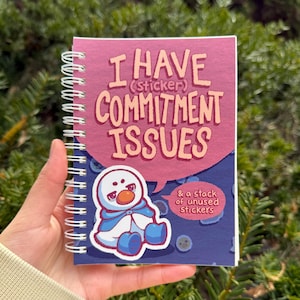 May include: A spiral-bound notebook with the text "I HAVE (sticker) COMMITMENT ISSUES" in pink and orange. A cartoon snowman sticker is on the bottom left. The notebook also says "& a stack of unused stickers".