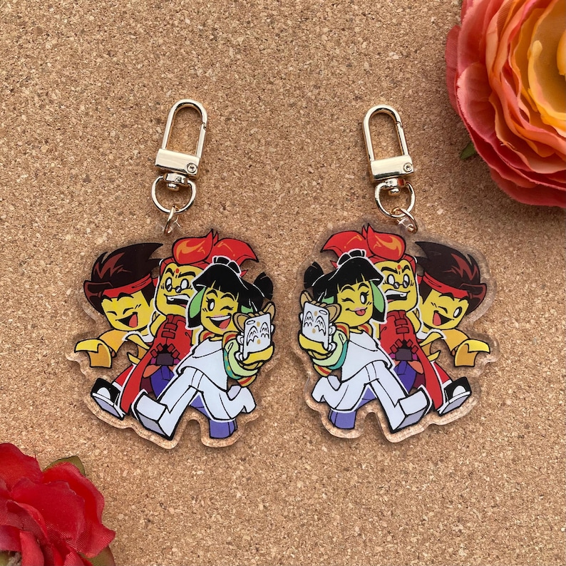 Puede incluir: Dos llaveros de acr&iacute;lico transparente con un grupo de personajes de dibujos animados. Cada llavero tiene un cierre dorado. Los personajes son de varios colores, incluyendo rojo, amarillo, negro y blanco. Los llaveros est&aacute;n sobre una superficie marr&oacute;n claro.