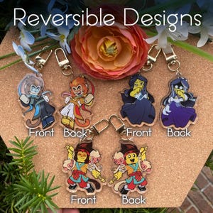 May include: Four reversible acrylic keychains featuring cartoon characters. The first keychain features a blue monkey with a yellow halo on the front and a yellow monkey with a red halo on the back. The second keychain features a purple character with a yellow halo on the front and a purple character with a blue halo on the back. The third keychain features a red monkey with a yellow halo on the front and a red monkey with a yellow halo on the back. The text "Reversible Designs" is at the top of the image.
