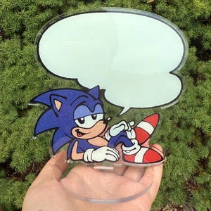 May include: A clear acrylic figure of Sonic the Hedgehog, in a relaxed pose, with a blank speech bubble. The character is blue with white gloves, red and white shoes, and a tan belly. The figure is held in front of a green, leafy background.