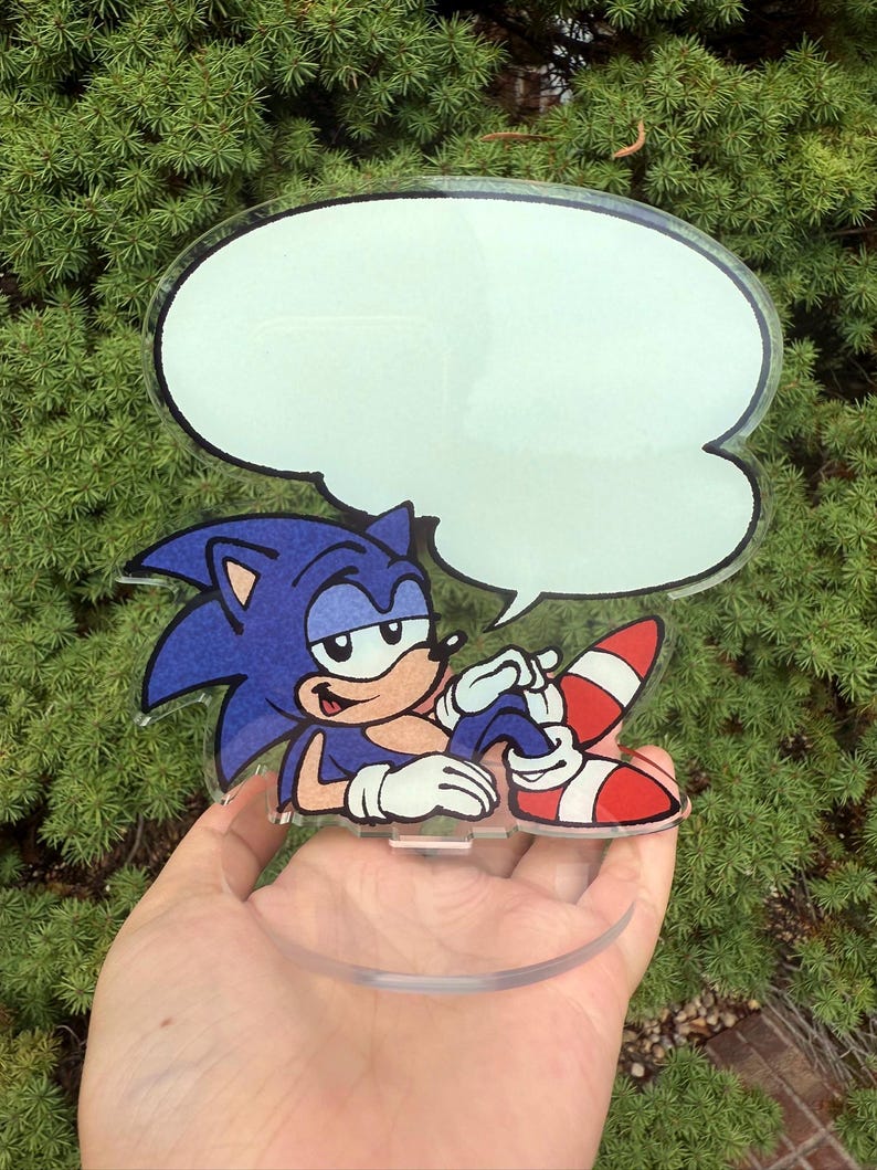 May include: A clear acrylic figure of Sonic the Hedgehog, in a relaxed pose, with a blank speech bubble. The character is blue with white gloves, red and white shoes, and a tan belly. The figure is held in front of a green, leafy background.
