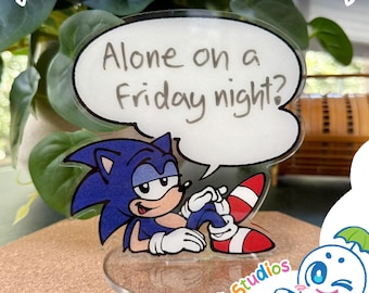 May include: A clear acrylic whiteboard standee featuring a graphic of the blue character Sonic the Hedgehog reclining. Above the character is a white speech bubble with the black text, Alone on a Friday night? The standee has a circular base.