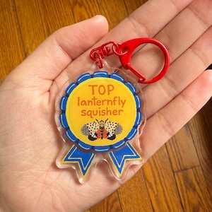 May include: A clear acrylic keychain with a red clasp. The keychain features a yellow circle with blue trim and the words "TOP lanternfly squisher". Below the text is an illustration of a lanternfly. Blue ribbons are at the bottom.