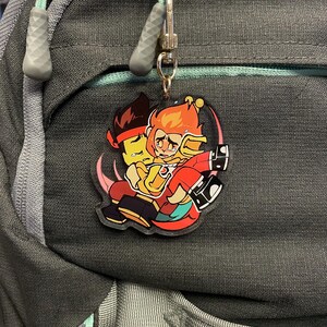 Puede incluir: Un llavero acr&iacute;lico colorido con dos personajes de dibujos animados abraz&aacute;ndose. Uno tiene pelo naranja y un atuendo rojo, el otro tiene pelo negro y una diadema roja. El llavero est&aacute; sujeto a una mochila.