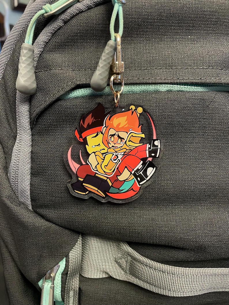 Puede incluir: Un llavero acr&iacute;lico colorido con dos personajes de dibujos animados abraz&aacute;ndose. Uno tiene pelo naranja y un atuendo rojo, el otro tiene pelo negro y una diadema roja. El llavero est&aacute; sujeto a una mochila.