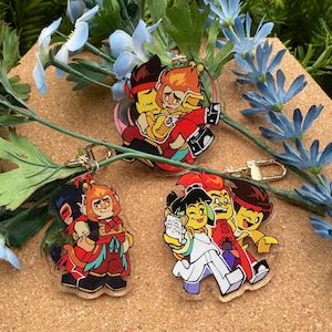 Puede incluir: Tres llaveros acr&iacute;licos con personajes de dibujos animados en varias poses. Los llaveros tienen cierres dorados. Los personajes est&aacute;n representados con trajes rojos, amarillos, negros y blancos. Los llaveros est&aacute;n sobre una superficie marr&oacute;n con detalles florales azules y verdes.