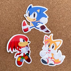 May include: Three colorful cartoon character stickers featuring Sonic the Hedgehog, Tails, and Knuckles. The stickers are on a brown corkboard background.