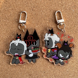 May include: Four colorful acrylic keychains featuring cartoon characters. A black cat with three eyes is wearing a maid outfit and holding a plate of food. A gray sheep with a red cape is holding a glass of wine. A gray sheep with a crown is wearing a red cape and holding a gold coin. A black and purple demon with horns is wearing a purple cape and holding a skull.