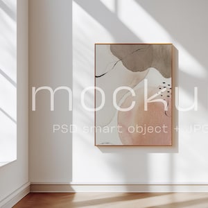 May include: A framed art print with a watercolor-style abstract design in neutral tones of peach, brown, and beige hangs on a white wall. The print is in a gold frame. The word "mockup" is printed on the wall. The room has a window and hardwood floor.