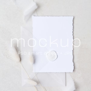 May include: A white envelope with a white card inside. The card has a white wax seal on it. There are two white pampas grass stems in the background.