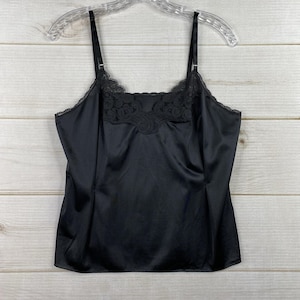 Op de afbeelding: Zwarte camisole met verstelbare bandjes en kanten details langs de halslijn. De camisole is gemaakt van een glad, satijnachtig materiaal en hangt aan een doorzichtige plastic hanger.