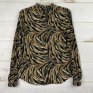 Pode incluir: Camisa de botão de manga comprida com estampa de zebra em preto, castanho e creme. A camisa tem gola e está pendurada em um cabide em um fundo de madeira clara.