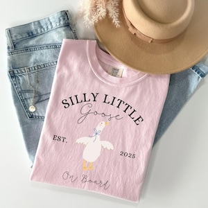 以下が含まれることがあります： 「Silly Little Goose」の文字とガチョウのイラストがプリントされた薄いピンク色のTシャツ。青いジーンズとベージュの帽子の上に置かれています。「Est. 2025 On Board」の文字もプリントされています。