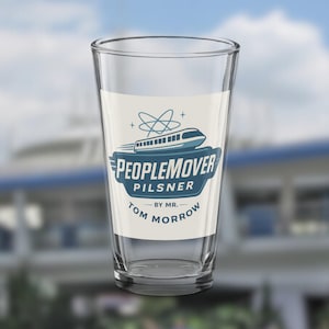Könnte beinhalten: Ein klares Pilsner-Glas mit einem weißen Etikett mit der Aufschrift "PeopleMover Pilsner" in Blau, zusammen mit einer stilisierten Zuggrafik. Der Text "by Mr. Tom Morrow" ist ebenfalls vorhanden. Das Glas ist leer.