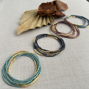 May include: Five beaded bracelets in different colors, including teal, blue, and purple, with gold accents. The bracelets are layered on a light gray surface.