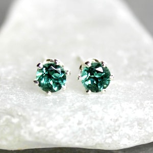 May include: A pair of stud earrings featuring round, faceted, teal-green gemstones set in silver. The earrings are displayed on a white, textured surface against a dark background. The gemstones have a brilliant cut.