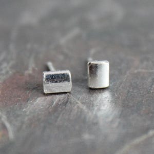 May include: A pair of small, silver-colored stud earrings. Each earring has a rectangular, rounded shape. The earrings are set against a textured, gray background.