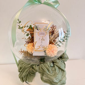 Peut inclure: Un ballon transparent rempli de fleurs séchées et de verdure, une boîte de chocolats Ferrero Rocher et un nœud en tissu vert. Le ballon est posé sur une base blanche avec un tissu vert autour.