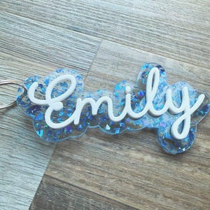 May include: A clear acrylic keychain with the name "Emily" in white script. The keychain has a cloud-like shape and is filled with blue glitter. A silver keyring is attached to the side.