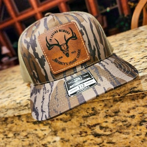 Op de afbeelding: Een camouflage trucker-pet met een bruin leren patch waarop staat "In Loving Memory 1948 - 2023 Searcy's Osage Lodge". De patch heeft een schedel en geweien. De pet is gemaakt door Richardson 112.