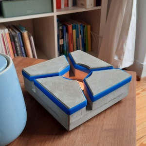 May include: A grey concrete candle holder with a geometric design. The holder has four sections with blue trim, surrounding a central space for a lit candle. The candle holder sits on a wooden surface.