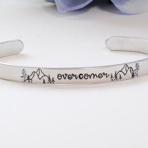 May include: Silver bracelet with the word "overcomer" engraved on it, along with a mountain range and trees on either side.