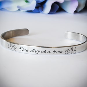 May include: Silver bangle bracelet with the inscription "One day at a time" and two small flower designs.