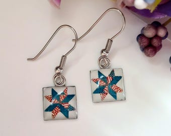 May include: A pair of square earrings with a patriotic star design. The earrings feature a silver hook and a square pendant with a star pattern in red, white, and blue, reminiscent of the American flag.