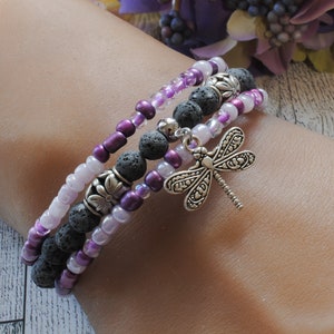 May include: A layered bracelet with purple and white beads, black lava rock beads, and a silver dragonfly charm.