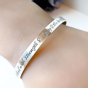 May include: A silver-toned metal cuff bracelet featuring a polished finish and engraved black script text that reads, "clothed with strength & dignity & she laughs". The bracelet has a simple, open-ended design that wraps around the wrist.