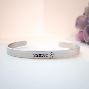 May include: A silver-colored cuff bracelet with the word "RESIST" and a fist symbol engraved on the front. The bracelet has a simple, open design and is likely made of metal. The background is a soft, neutral color.